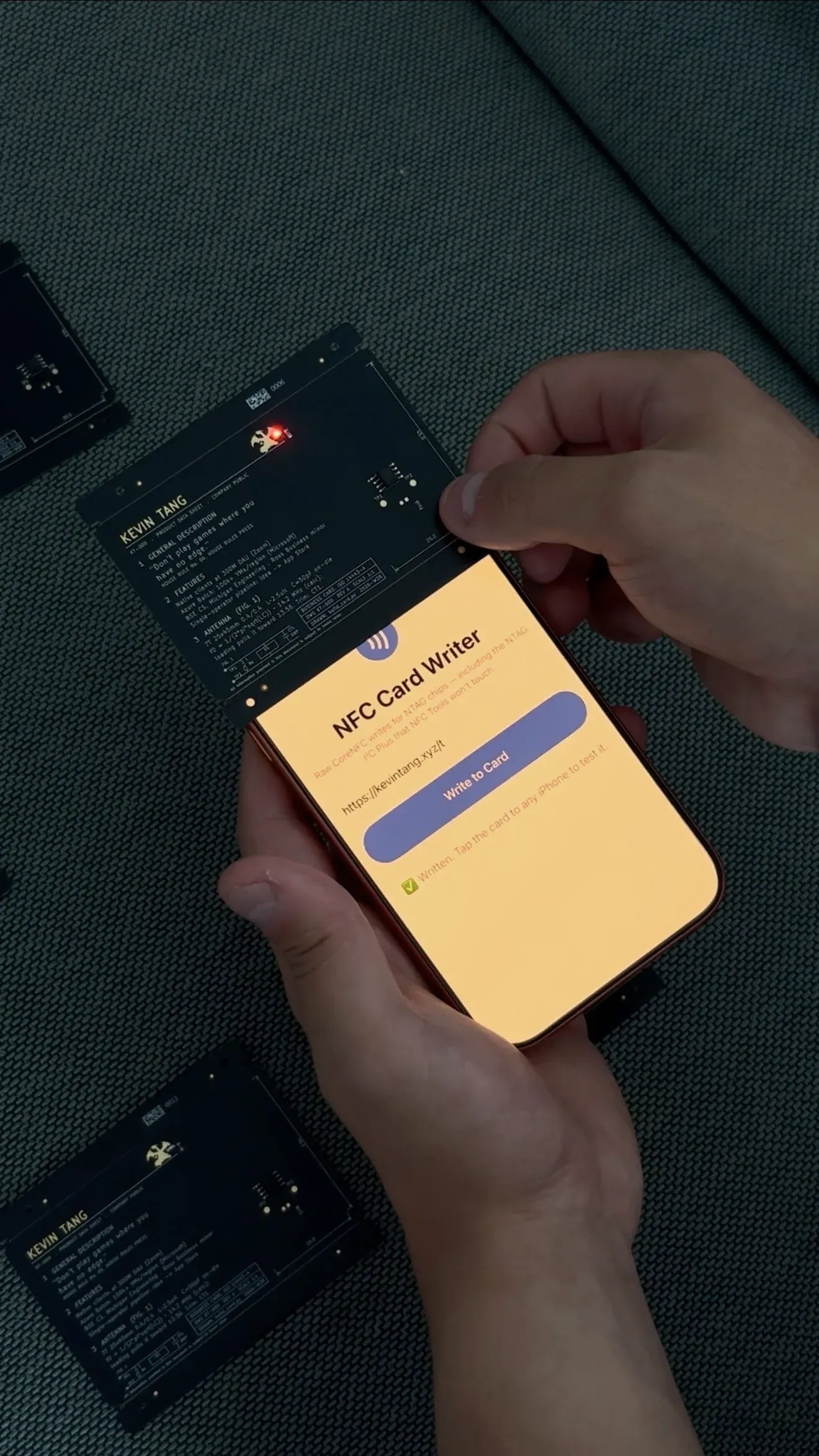 A black circuit-board business card styled like a component datasheet, a small red LED glowing near the top; below it a phone runs a custom "NFC Card Writer" app showing the URL being written to the card, with two more cards resting on a dark couch.