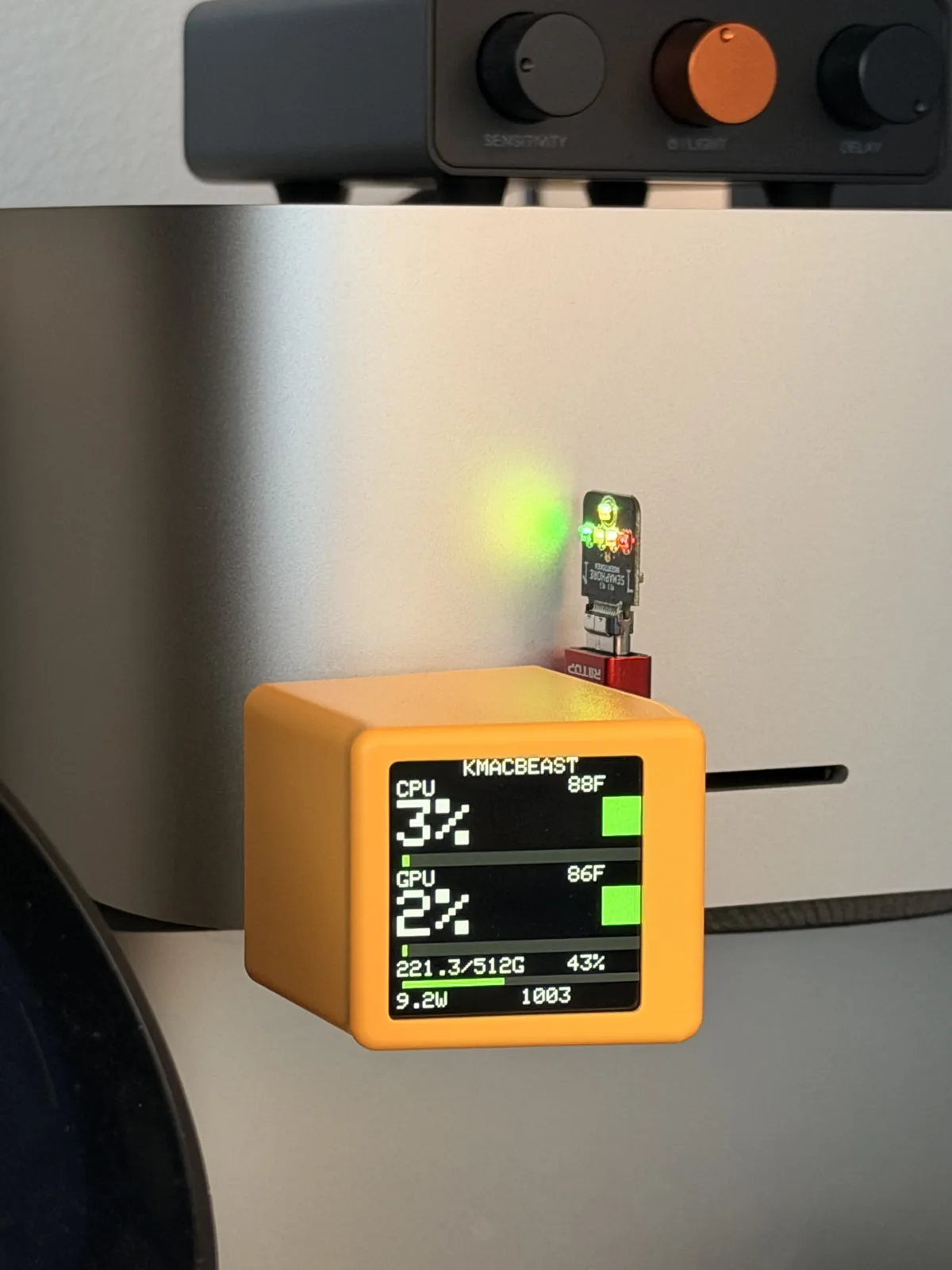 A Semaphore board with green, amber, and red LEDs plugged into a Mac Studio's front USB-C port, just above an orange SmallTV cube showing the KMACBEAST stats