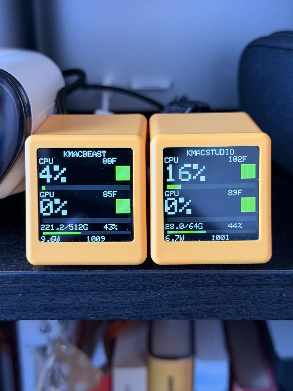 Two orange SmallTV cubes on a shelf, one labeled KMACBEAST and one KMACSTUDIO, each showing CPU and GPU percent, temperatures, memory, watts, and fan RPM